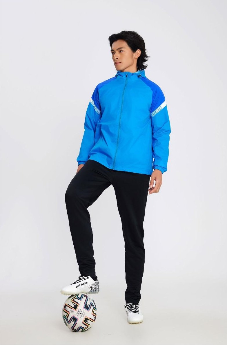 Man wearing blue hooded soccer windbreaker with black pants and football under foot