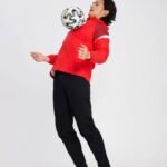Man in red soccer windbreaker balancing football on head in dynamic pose