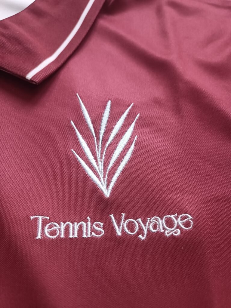 Close-up of high-density white direct embroidery Tennis Voyage brand logo on maroon pique mesh polo shirt.