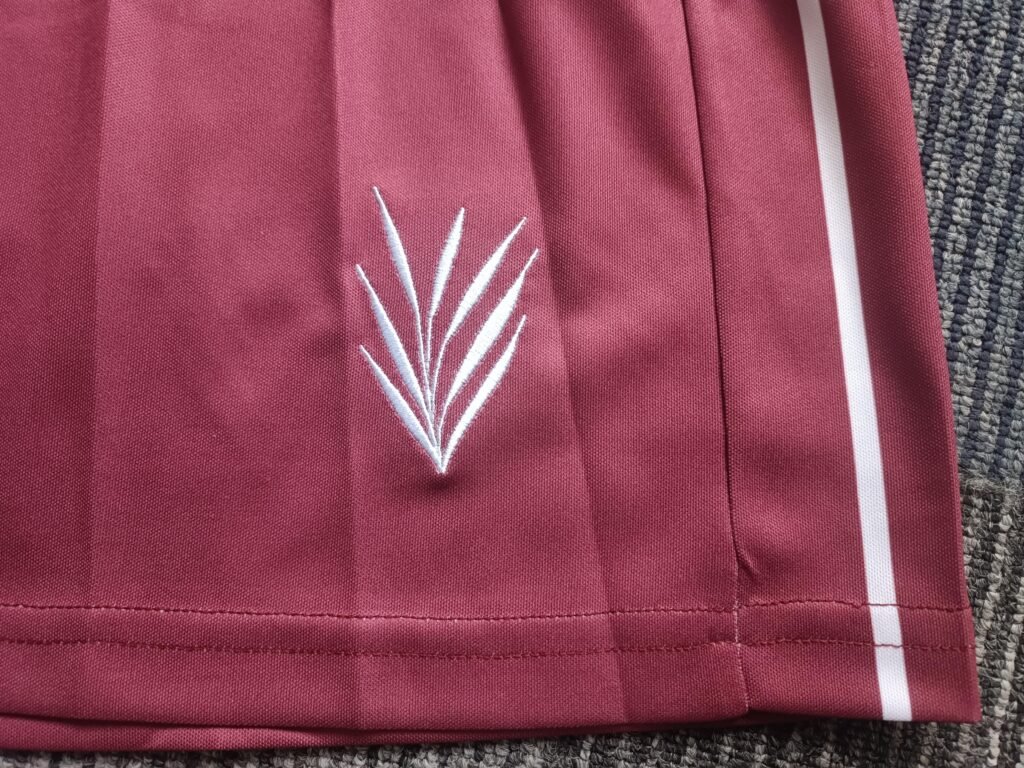 Detailed view of high-density white embroidered leaf emblem on the hem of maroon sublimated tennis shorts with side stripe.