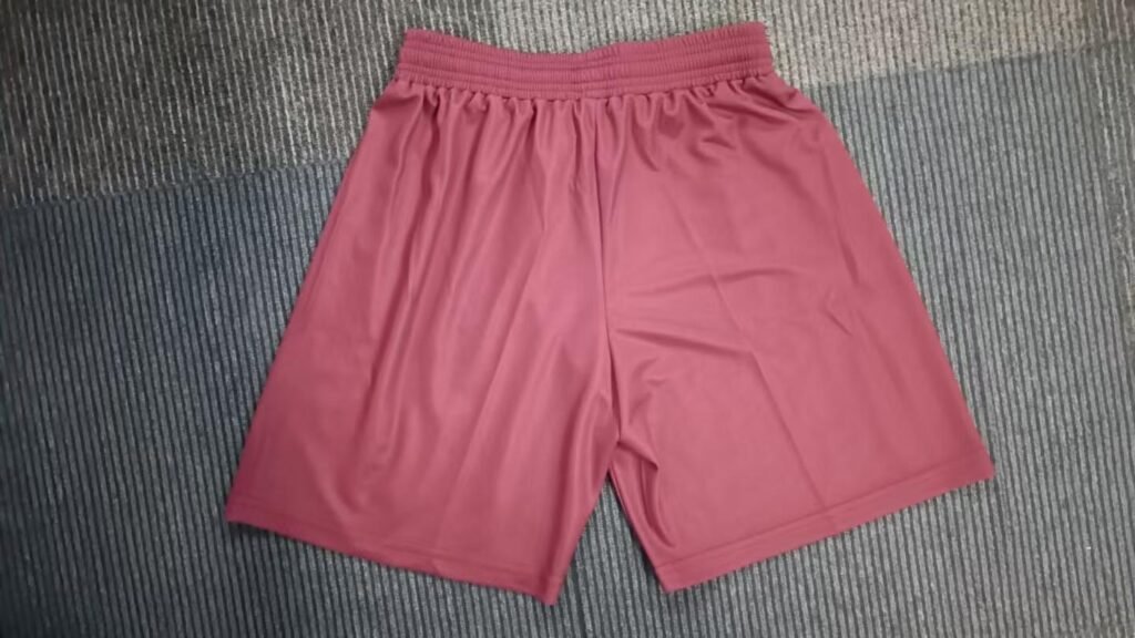 Flat-lay view of matching maroon athletic tennis shorts with white drawstrings, side seam stripes, and embroidered logo on left leg.
